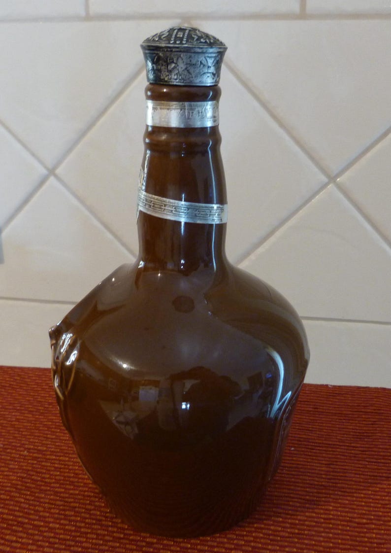 May include: A brown ceramic bottle with a silver top and a silver band around the neck. The bottle has a unique shape with a handle on the side.