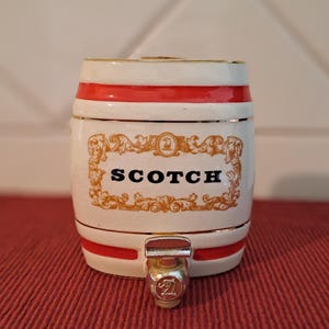 May include: A ceramic barrel-shaped container with a spigot, featuring a white base with red bands and gold trim. The word "SCOTCH" is displayed in black letters on a gold decorative label. The container is designed to hold and dispense scotch.