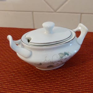 以下が含まれることがあります： ふた付きの白い磁器製の砂糖入れ。ふたには銀色の縁取りがあり、砂糖入れには緑の葉が描かれた花柄が施されています。