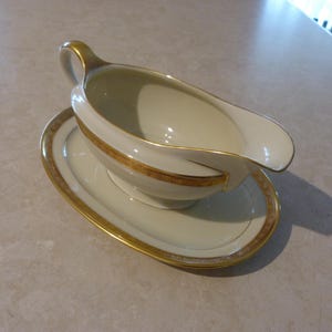 May include: A cream-coloured gravy boat and matching oval saucer. The boat has a curved handle and a pouring spout. Both pieces are trimmed with gold detailing, adding a touch of elegance to the serving set. The set is suitable for formal dining.