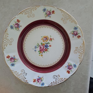 Op de afbeelding: Een witte porseleinen bord met een geschulpte rand, met een bordeauxrood midden en een bloemenontwerp. Het bord is versierd met gouden filigraan en kleine clusters van kleurrijke bloemen, waaronder roze, blauwe en gele bloemen.