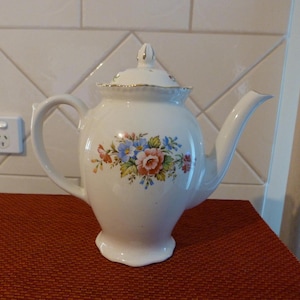 May include: White porcelain teapot with a floral design featuring pink, blue, and yellow flowers. The teapot has a gold trim around the lid and spout.