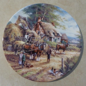 May include: A vintage style painting of a rural scene with a cottage, hay wagon, horses, and people. The painting is on a white ceramic plate with a gold rim.