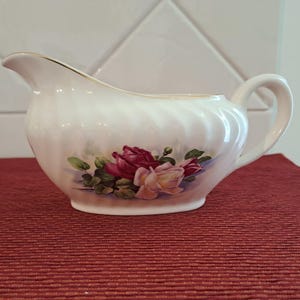 Op de afbeelding: Een witte keramische juskom met een gouden rand en een handvat. De kom heeft een bloemenontwerp van rode en roze rozen met groene bladeren. De juskom staat op een rode ondergrond.