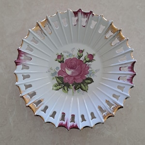 May include: White porcelain decorative bowl with a floral rose design. The bowl has a scalloped edge with gold trim and burgundy accents. The center features a large pink rose with green leaves and buds. The bowl has a radial design.
