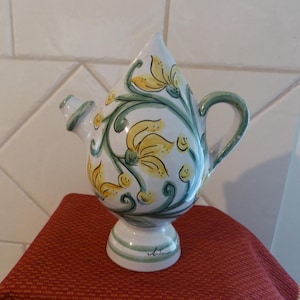 May include: A white ceramic pitcher with a teardrop shape, adorned with yellow floral designs and green vines. The pitcher has a small spout, a curved handle, and sits on a pedestal base. The base is decorated with green stripes.