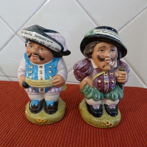 May include: Two ceramic figurines of men wearing hats and colorful clothing. The figurine on the left has a blue and white outfit and a black hat with the text "Burston Potteries £2.99 a better gift - call in today". The figurine on the right has a purple and green outfit and a black hat with the text "A favourite amongst those that know their real ale".