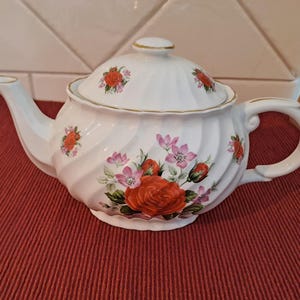 以下が含まれることがあります： 赤いバラとピンクの花柄と金色のトリムで飾られた、蓋付きの白い磁器のティーポット。 ティーポットは、曲がったハンドルと注ぎ口を備えています。 クラシックなティーアクセサリーです。