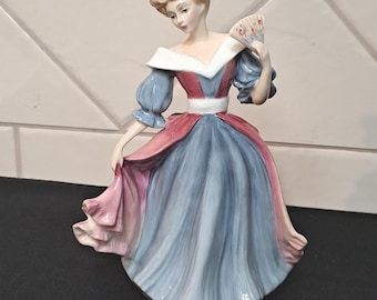 May include: A porcelain figurine of a person with blonde hair styled in an updo, wearing a dress with a white off-the-shoulder neckline, blue puffed sleeves, and a skirt featuring alternating panels of dusty blue and pink, holding a small hand fan.