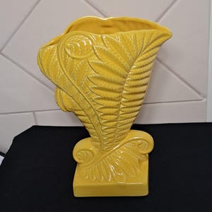 May include: A bright yellow ceramic vase featuring a raised, textured fern leaf pattern that wraps around the body. The vase has a wide, flared opening at the top and sits on a square base, with curled leaf details accenting the lower section.