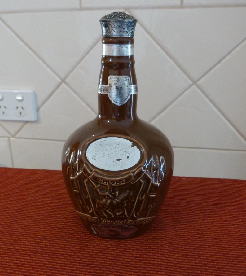 May include: A brown ceramic bottle with a silver top. The bottle has a label that says "Chivas Regal" and a crest with a knight on a horse. The bottle is sitting on a red surface.