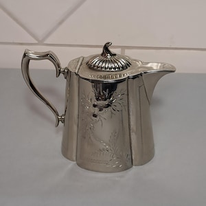 May include: A polished silver-tone metal teapot featuring a hinged lid with a decorative finial, a curved handle, and a short spout. The body of the teapot is adorned with an engraved floral pattern consisting of delicate leaves and blossoms.