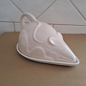 Peut inclure: Un beurrier en céramique rose clair en forme de souris. Le plat a une poignée incurvée et repose sur une base ovale blanche. Les traits de la souris sont subtilement détaillés, avec une finition lisse et brillante.