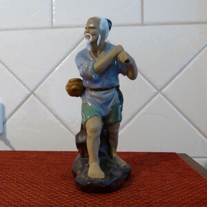 May include: A ceramic figurine of an elderly man in traditional attire. The figure has a white beard and hair, wearing a blue tunic, green shorts, and holding a small brown pot. The figurine is mounted on a dark base.