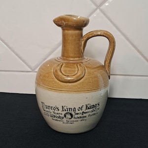 May include: A two-toned ceramic whiskey jug with a tan upper section and a cream lower section. It features a handle, a spout, and a horseshoe-shaped relief. The text reads: Munro's King of Kings, Twenty Years Old Whisky, James Munro & Son Ltd, Dalwhinnie Distillery, Strathspey, Inverness-shire, Scotland.