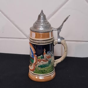 May include: A ceramic beer stein featuring a dark blue body with a colourful relief scene of a village and mountains. It has a tan base and rim, a curved handle, and a pointed, hinged metal lid with intricate embossed detailing on the surface.