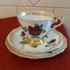 May include: A white teacup, saucer, and side plate set with a red rose design and gold trim. The teacup has a gold handle and base. The set is on a red surface.