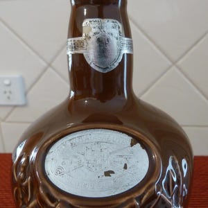 May include: A brown ceramic bottle with a white label that says "Royal Salute" and a silver label with a crest. The bottle is shaped like a traditional whisky bottle.