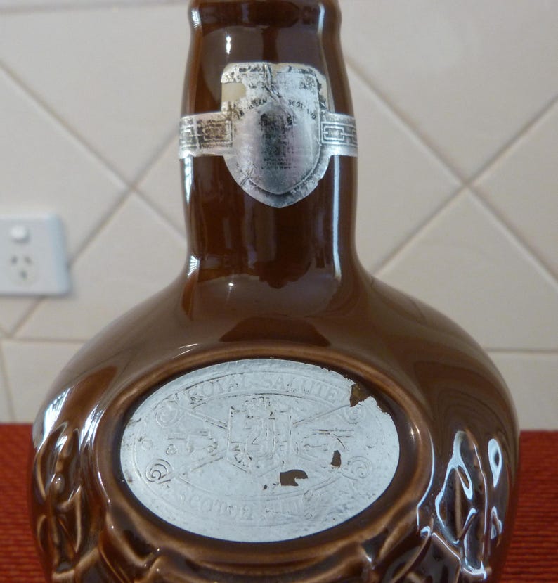 May include: A brown ceramic bottle with a white label that says "Royal Salute" and a silver label with a crest. The bottle is shaped like a traditional whisky bottle.