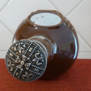 May include: A brown ceramic bottle with a silver metal top. The top features a decorative design with a cross and fleur-de-lis.