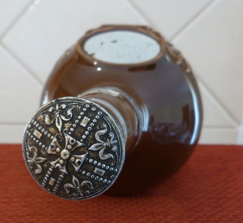 May include: A brown ceramic bottle with a silver metal top. The top features a decorative design with a cross and fleur-de-lis.