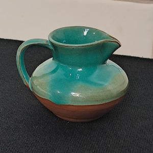 May include: A small ceramic jug with a rounded body and a curved handle. The upper portion features a glossy, vibrant turquoise glaze that drips down towards the base, while the lower section remains an exposed, matte brown clay finish.