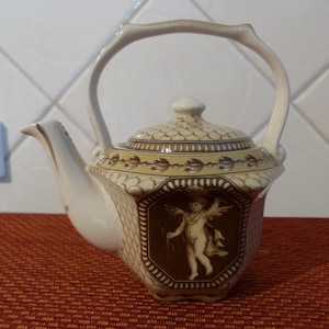 May include: Cream ceramic teapot with a decorative handle and spout. The teapot has a brown and cream design, featuring a cherub image. The lid has a matching pattern. The teapot is on a red surface. The teapot is approximately 20 cm in height.
