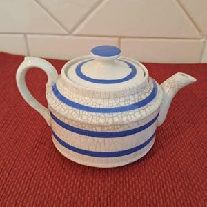 May include: A white ceramic teapot with blue stripes and a matching lid. The teapot has a curved handle and a spout. The design features a crackle glaze effect. The teapot is sitting on a red surface.