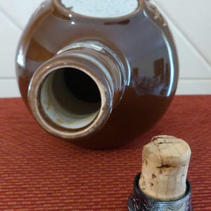 May include: A brown ceramic bottle with a silver-coloured rim and a cork stopper. The bottle has a white label on the top.
