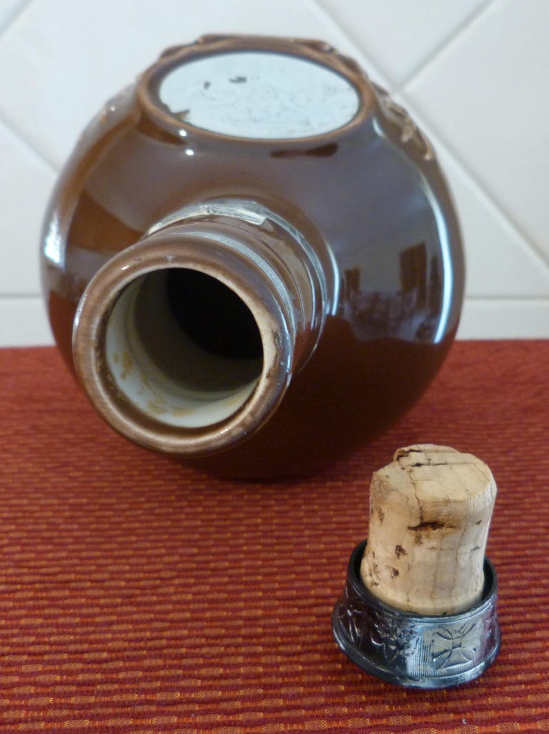 May include: A brown ceramic bottle with a silver-coloured rim and a cork stopper. The bottle has a white label on the top.