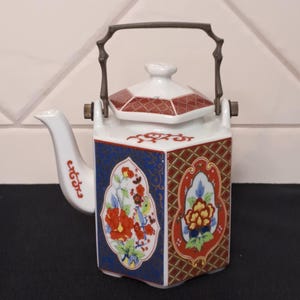Può includere: Una teiera esagonale in porcellana con base bianca e pannelli alternati di motivi geometrici blu scuro e rosso ruggine. Ogni pannello presenta intricati motivi floreali in rosso, giallo e verde. Dotata di manico in metallo e coperchio.