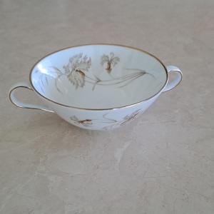 May include: White ceramic soup bowl with two handles and a gold rim. The bowl features a floral design in shades of brown and grey. A delicate and elegant piece for serving soup or other dishes.