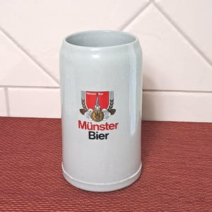 May include: A light grey ceramic beer stein with the words "Münster Bier" in red and black. The stein features a crest with a red background and wheat stalks. The cylindrical stein has a handle.