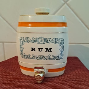 May include: A white ceramic rum barrel with orange bands and gold trim. The barrel features a decorative blue and white label with the word "RUM" in black. It has a spigot and a cork stopper.