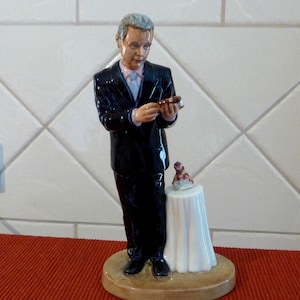 以下が含まれることがあります： 黒いスーツを着た男性の陶器製の置物で、葉巻を持っています。丸い台座の上に立っており、小さなテーブルと装飾品がそばにあります。置物は顔が細かく、白いタイルの背景に配置されています。