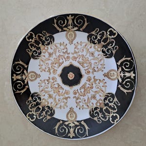 May include: A round decorative plate with a black and white design. The plate features a black border with gold scrollwork and a white center with gold floral patterns. The plate has a gold rim.