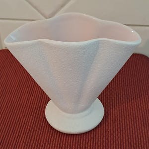 May include: A white, textured vase with a flared, scalloped rim. The vase has a conical body and a round base. The interior is a soft pink. The vase is likely ceramic and suitable for floral arrangements or decorative use.