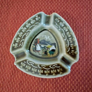May include: A triangular ashtray with a green-gray glaze. The center features a landscape scene with the word "Ireland". The rim is decorated with shamrocks. The ashtray has three indentations for cigarettes.