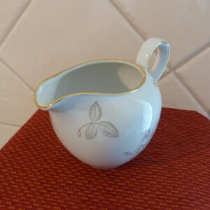 May include: A white ceramic creamer with a gold rim and handle. The creamer has a delicate grey leaf design. It is on a red-orange textured surface.