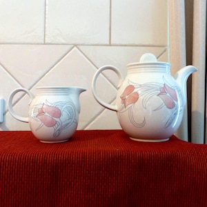 May include: A white teapot and creamer set with a floral design featuring pink tulips and gray leaves. The teapot has a lid and a handle. The creamer has a handle.