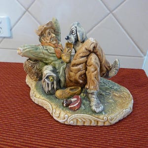 May include: A ceramic figurine of a man and his dog relaxing on a grassy patch. The man is wearing a hat, a green jacket, and brown pants. The dog is sitting next to him, looking at the viewer. The figurine is painted in realistic colors and has a detailed finish.