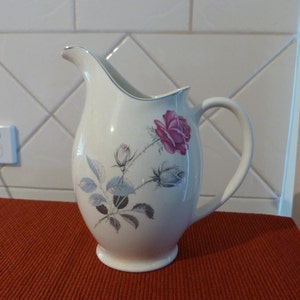 Op de afbeelding: Een witte keramische kan met een bloemenmotief met een roze roos en grijze bladeren. De kan heeft een brede opening en een handvat.