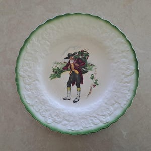 May include: A white decorative plate with a green scalloped edge. The plate features a raised floral pattern around the rim and a central illustration of a person carrying a barrel of grapes. The person is wearing a hat and traditional clothing.