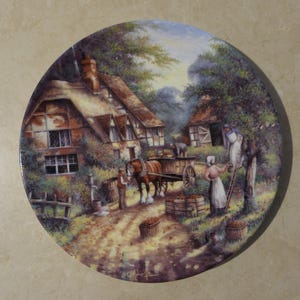 May include: A decorative plate featuring a pastoral scene with a horse-drawn cart, a cottage, and figures harvesting. The artwork depicts a rural landscape with a dirt road, trees, and a sunny sky. The plate has a glossy finish.