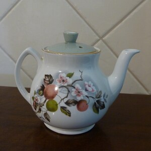 May include: A white teapot with a light blue lid and a floral design featuring pink flowers, green leaves, and orange fruit. The teapot has a gold trim around the lid and spout.
