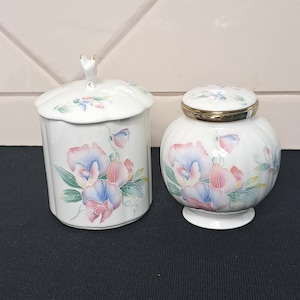 May include: Two white porcelain jars featuring a delicate floral pattern of pink, blue, and green sweet pea flowers. The taller jar has a scalloped lid with a small handle, while the shorter, rounder jar features a gold-toned metal rim on the lid.