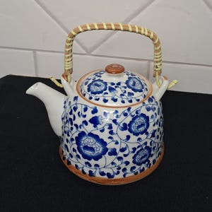 May include: A white ceramic teapot featuring a blue floral pattern with vines and leaves. The teapot has a matching lid with a brown knob, a curved spout, and a light-coloured woven bamboo handle attached to two upright loops on the top rim.