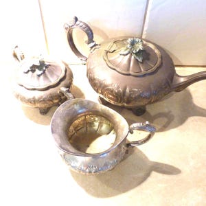 Op de afbeelding: Een zilveren theeservies met een theepot, roomkan en suikerpot. De set heeft een sierlijk ontwerp met bloemmotieven.