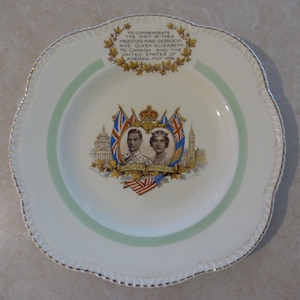 Op de afbeelding: Een wit keramiek bord met een groene en gouden rand. In het midden van het bord staat een herdenkingsbeeld van koning George VI en koningin Elizabeth, met de tekst "TO COMMEMORATE THE VISIT OF THEIR MAJESTIES-KING GEORGE VI AND QUEEN ELIZABETH TO CANADA AND THE UNITED STATES OF AMERICA MAY 1939" en "LONG MAY THEY REIGN".