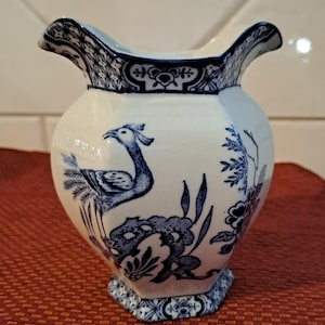 May include: A white and blue ceramic jug with a bird and floral pattern. The jug features a flared rim and a hexagonal base. The blue design is intricate and covers the entire surface of the jug.